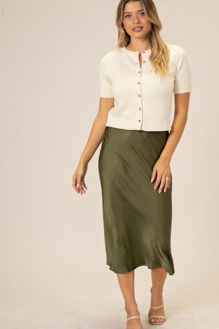 A woman wearing a cream-colored cardigan sweater with button-up front and a long olive green slip skirt, modeling the Ivana Satin Slip Skirt product.