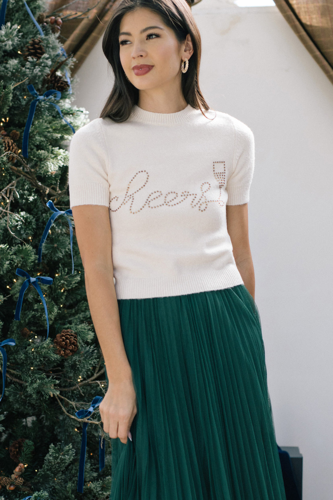 A white knitted sweater with the word "Cheers" spelled out in rhinestones, paired with a long pleated green skirt, modeled by a woman with long dark hair.