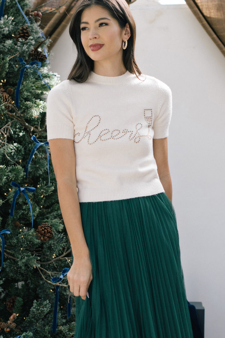 A white knitted sweater with the word "Cheers" spelled out in rhinestones, paired with a long pleated green skirt, modeled by a woman with long dark hair.