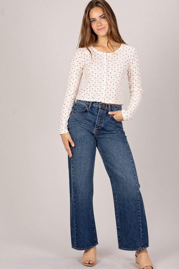Woman wearing a white knit top with pink floral print, paired with high-waisted blue denim jeans, posing against a neutral background.