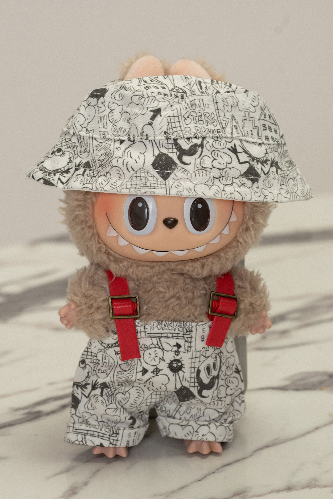 A plush stuffed animal with a fuzzy gray body, wearing a patterned overalls with a whimsical cartoon design, and a patterned bucket hat with large eyes.