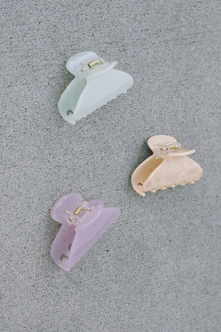 Three colored claw hair clips in pink, lilac, and white with gold-tone metal accents.