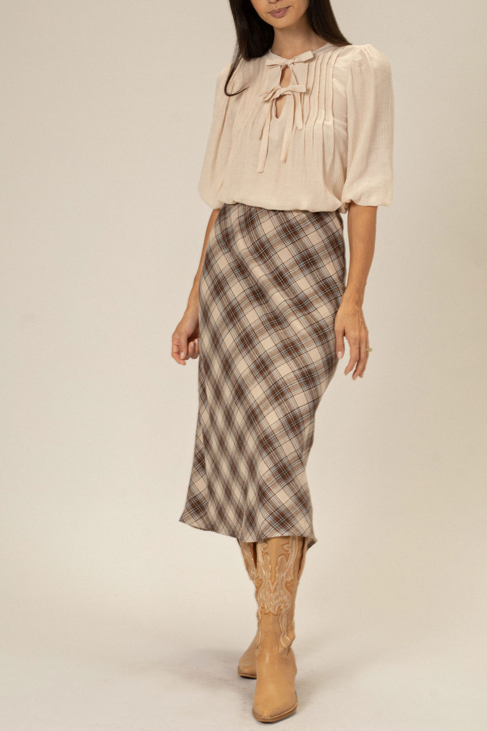 A beige blouse with puffed sleeves and a ribbon tie at the neck, paired with a brown and white plaid midi skirt and tan cowboy boots.