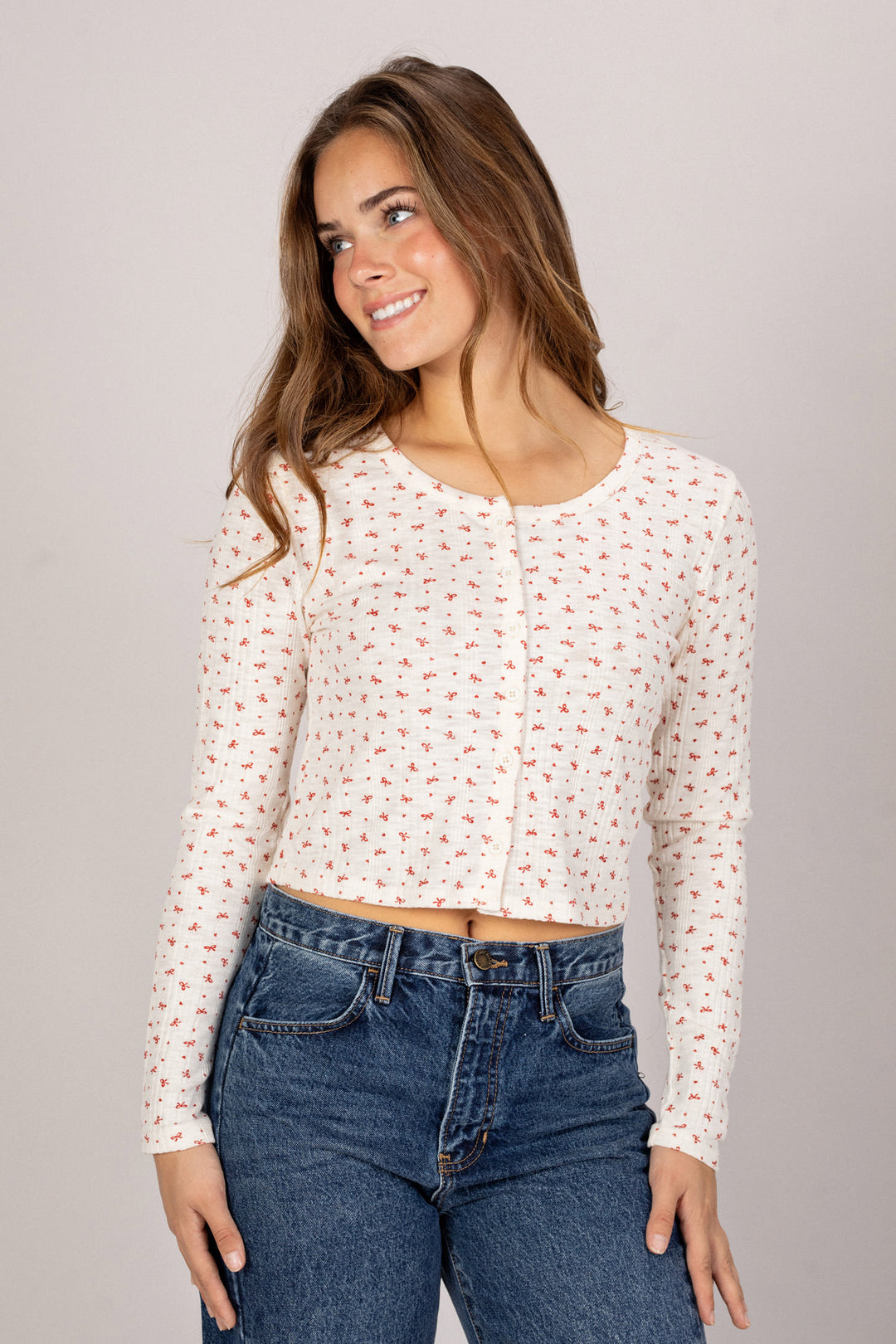 A young woman with long brown hair smiles while wearing a white knit top with small red floral print, paired with high-waisted blue jeans.