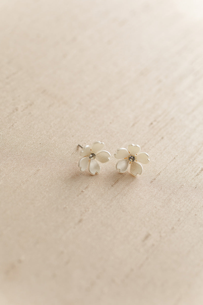 Two small white enamel flower stud earrings with crystal centers on a beige linen background.