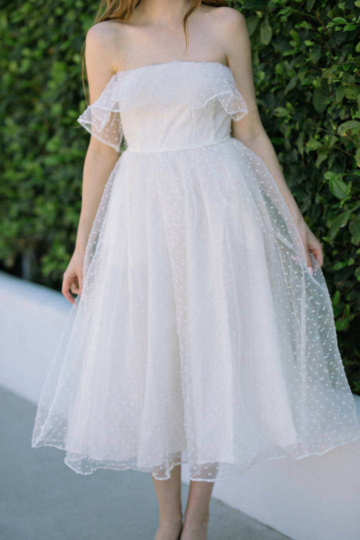 A white off-the-shoulder midi dress with a tulle overlay and polka dot pattern, featuring a voluminous skirt and flutter sleeves.