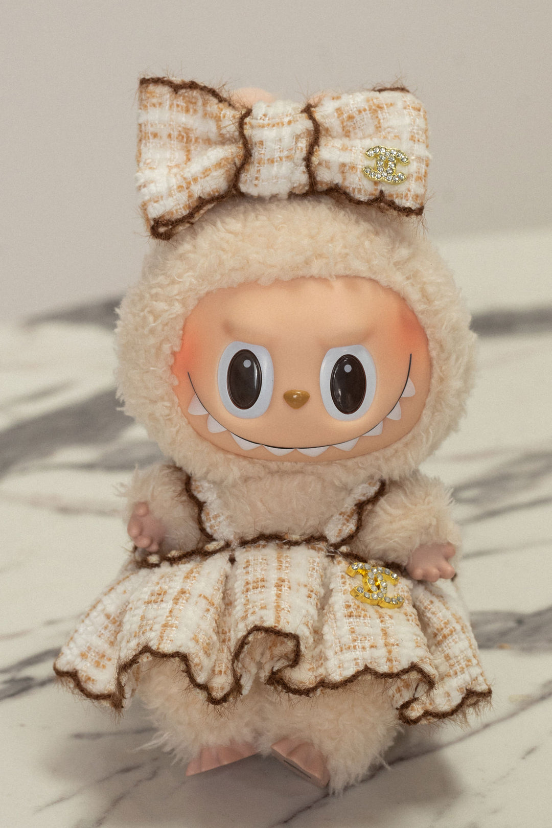 Soft, furry plush doll with large eyes, wearing a textured brown and beige tweed outfit and headband with a Chanel-inspired charm.