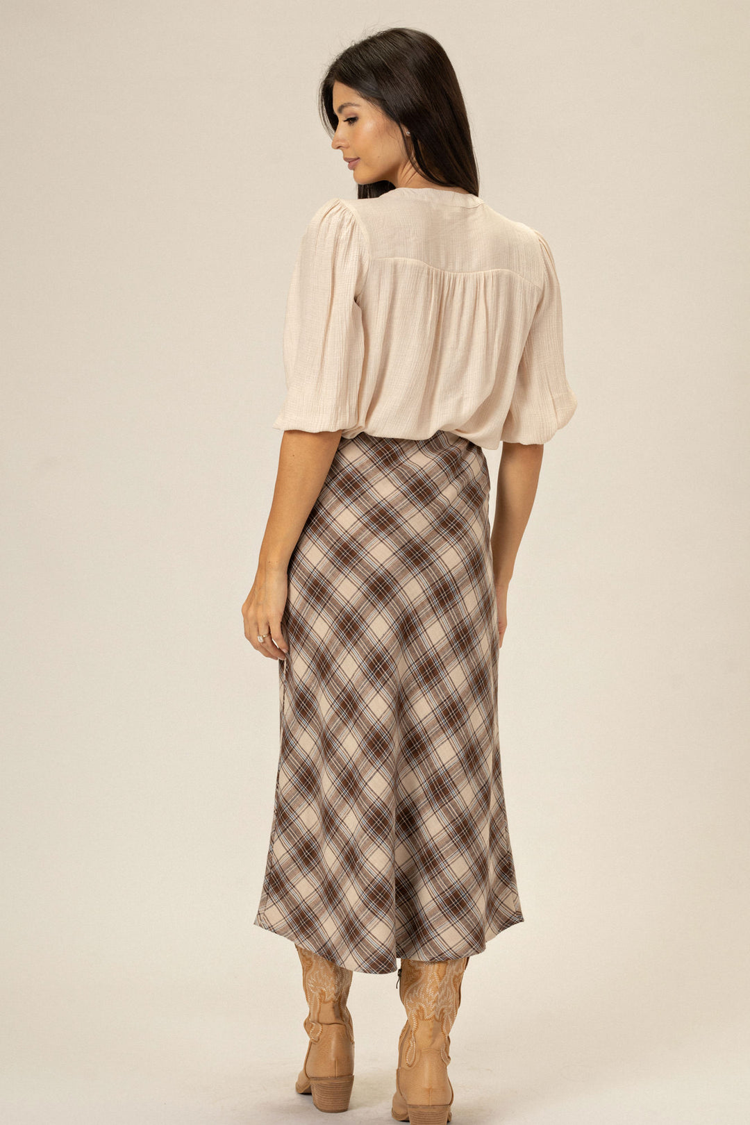 A light-colored, loose-fitting blouse with puffy sleeves, paired with a plaid midi skirt featuring a faint brown, beige, and white checkered pattern, worn with tan ankle boots.