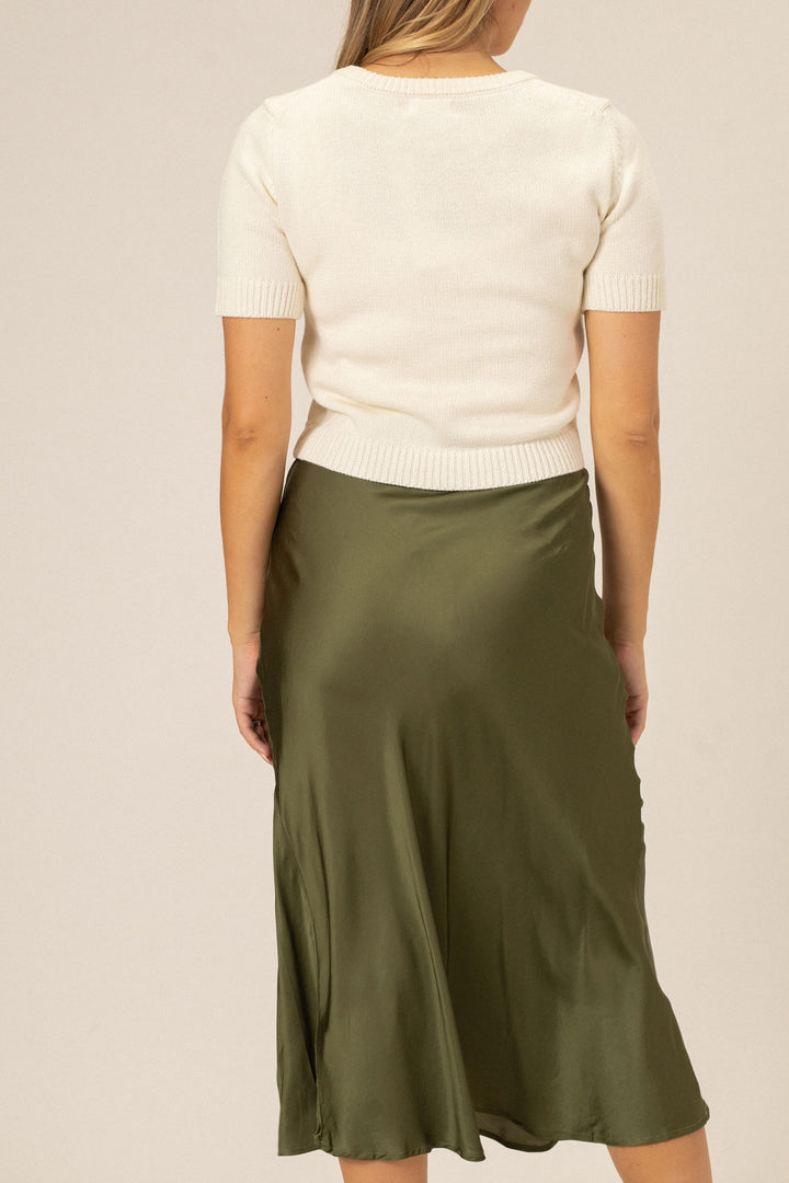 A cream-colored knit top with short sleeves and a high neckline, paired with a long olive green satin slip skirt.