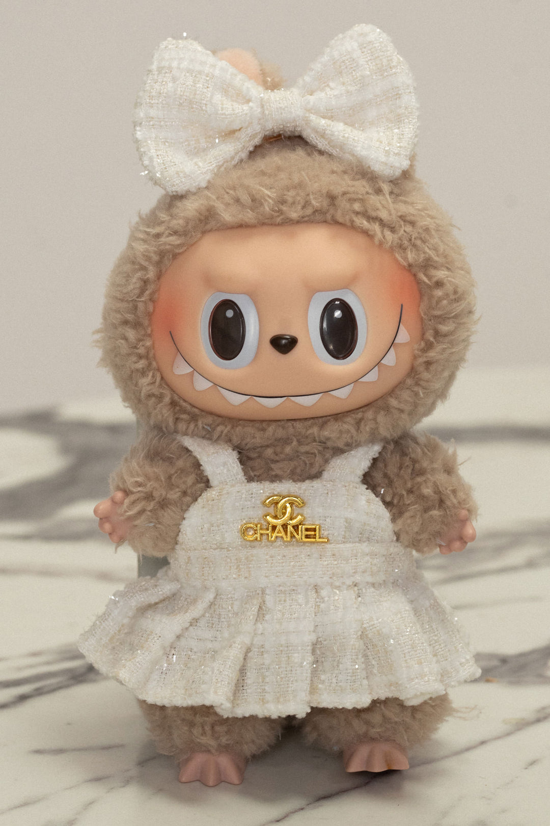 Plush toy sloth wearing a white tweed dress with a large white bow and a Chanel logo pin.