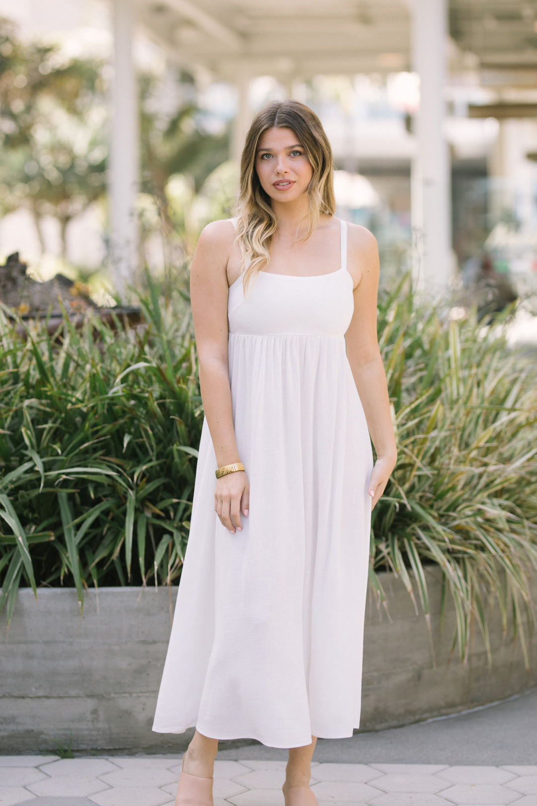 A young woman wearing a white, linen-blend midi dress with a relaxed, empire-waist silhouette, posing in a lush, garden-like setting.
