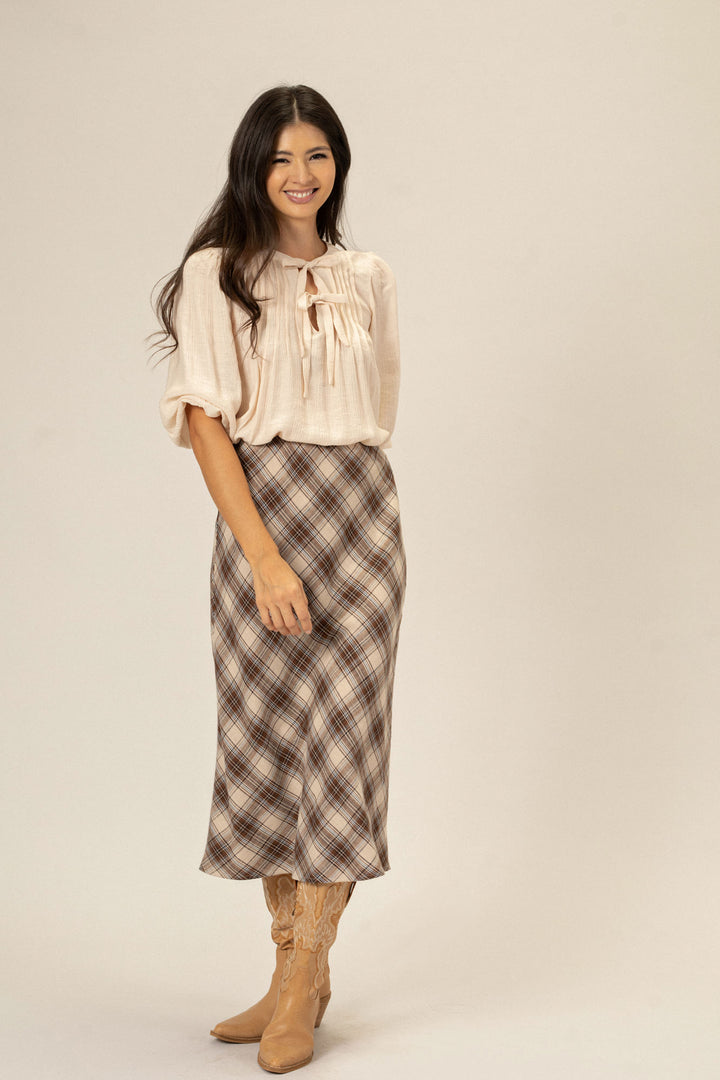 A woman wearing a cream-colored blouse with a bow neckline and a plaid midi skirt paired with tan cowboy boots, smiling at the camera.