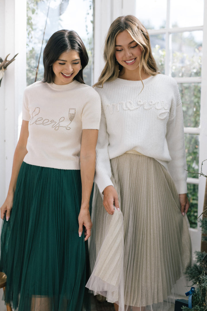Two women wearing soft, cream-colored sweaters with embellished text details, paired with pleated skirts in green and beige shades.