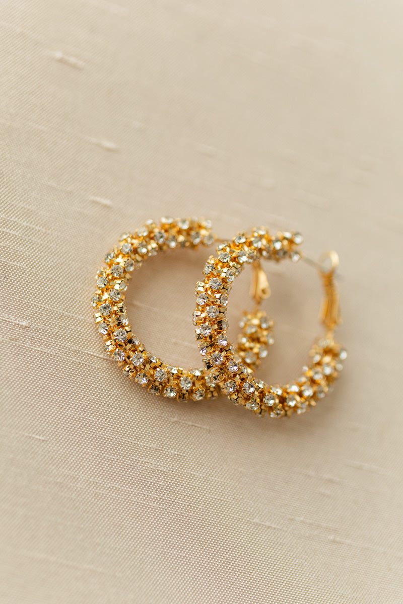 Sparkling gold-toned hoops adorned with glittering crystal stones, creating a glamorous, eye-catching statement accessory.