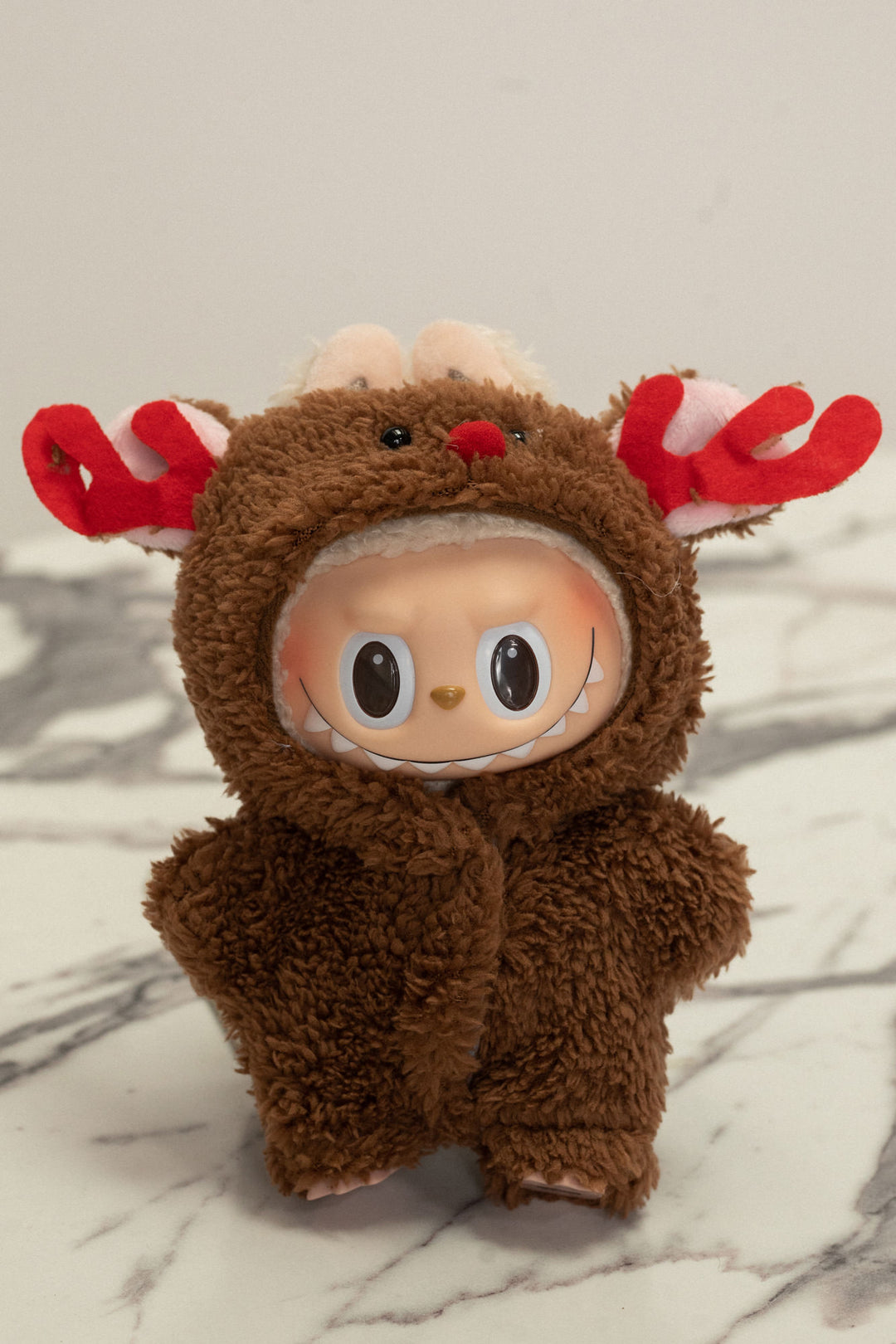 A plush brown stuffed animal with antlers, red nose, and big eyes wearing a cozy moose-themed jumpsuit.
