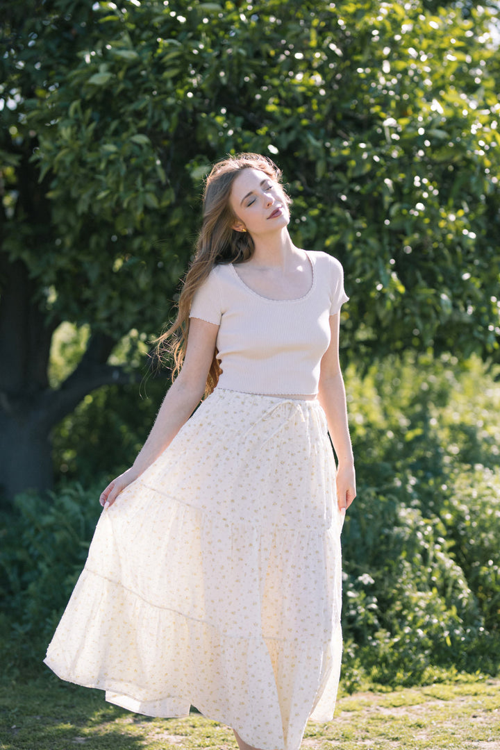 A young woman with long brown hair wearing a white cropped top and a long white floral skirt standing in a lush green garden.