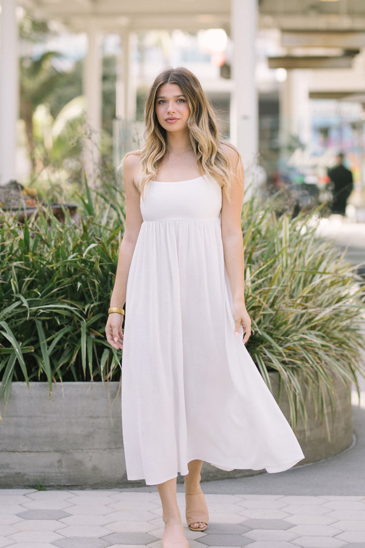 A young woman in a flowing, ivory midi dress with a fitted bodice and flared skirt stands in an outdoor, garden-like setting.