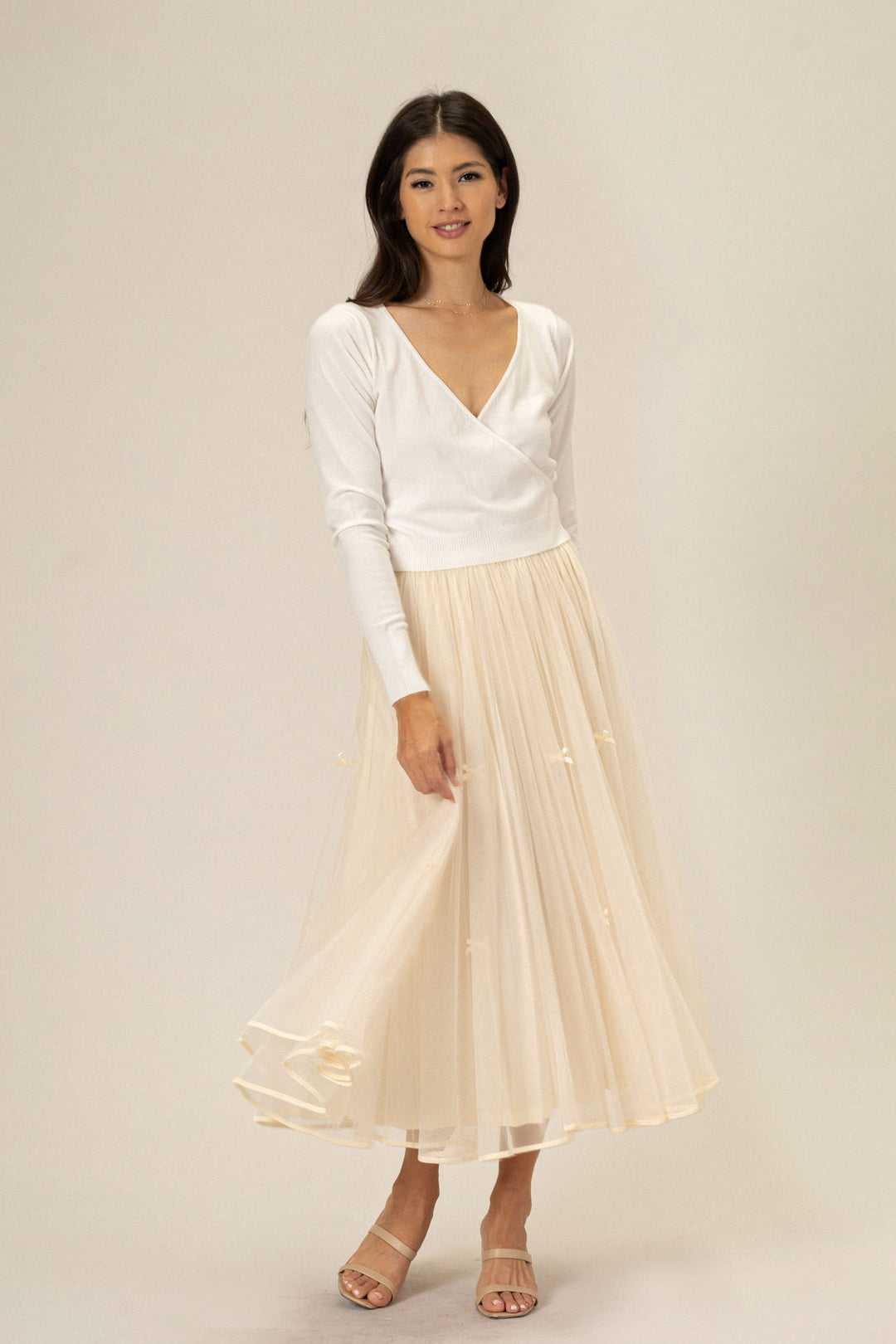A woman in a white wrap-style top and a long, flowing off-white tulle skirt with a bow detail, standing against a plain background.