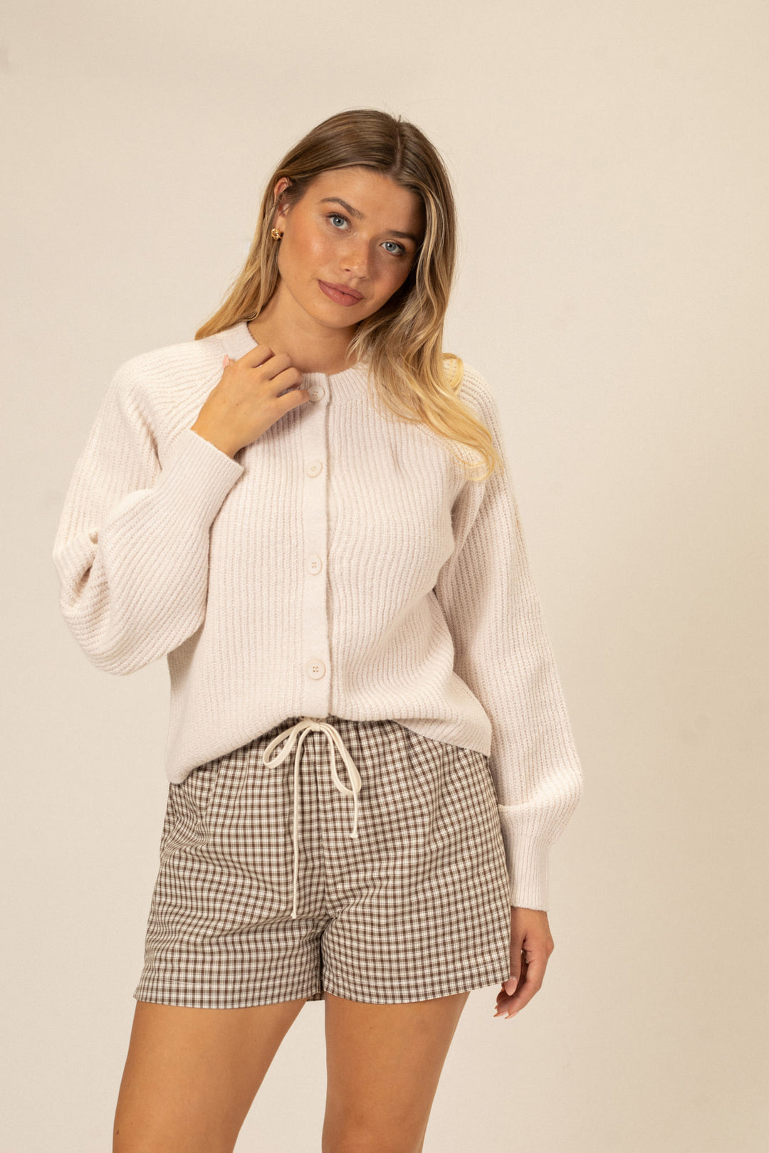 A woman wearing a white ribbed cardigan and plaid drawstring shorts stands against a beige background.