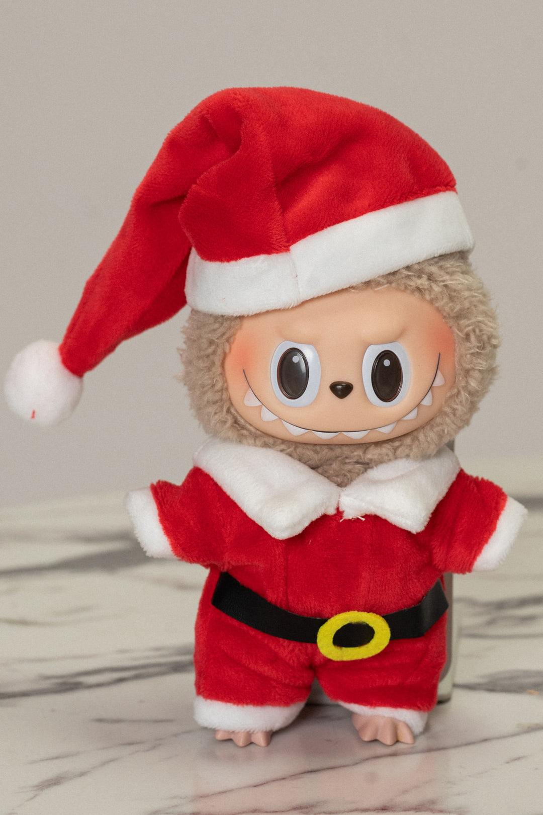 A soft, plush stuffed animal wearing a bright red Santa Claus suit with a white collar, belt, and boots. It has a smiling expression and large black eyes.