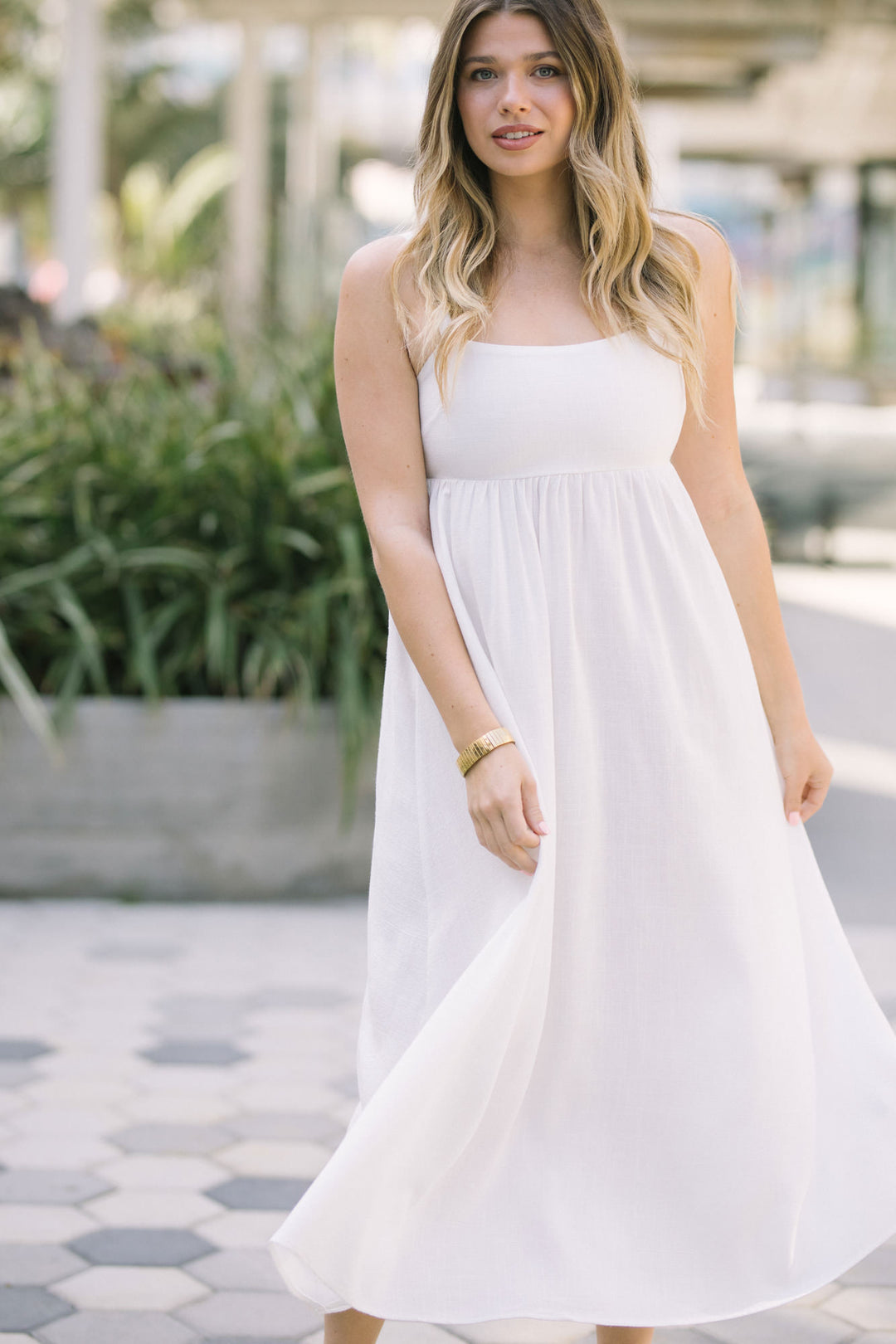 A woman wearing a flowy, white midi dress with a sleeveless, A-line silhouette, standing in a tropical setting surrounded by greenery.