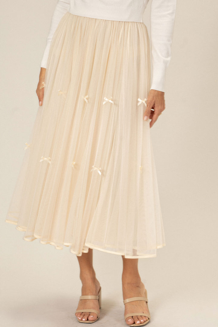A full, flowing ivory tulle skirt featuring delicate white bows cascading down the front, paired with beige slip-on sandals.