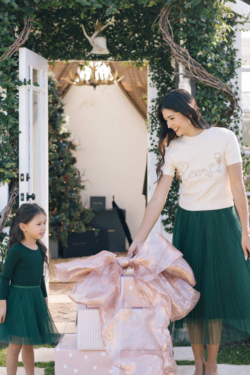Two women, one adult and one child, stand in an outdoor setting. The adult wears a white top with the text "Cheers" and a pleated green skirt. The child wears a green dress. They are surrounded by greenery and an arched doorway.