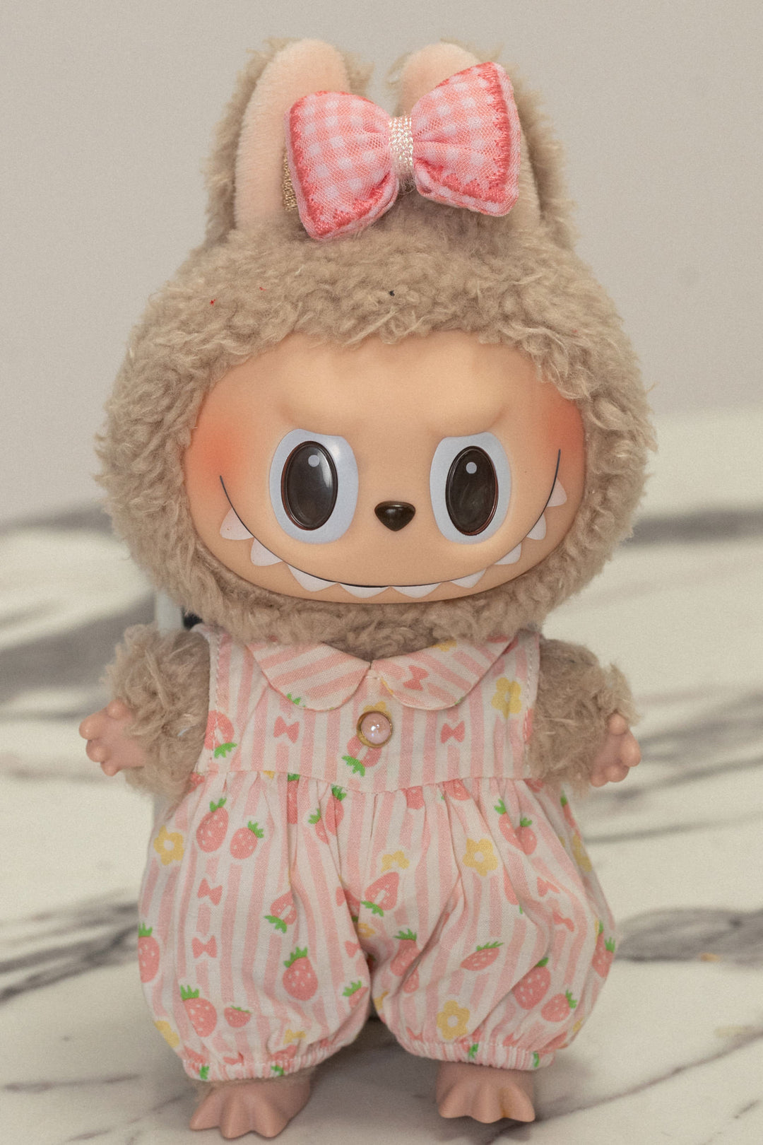 A plush, gray toy with large eyes and a pink bow, wearing a patterned pink jumpsuit with a strawberry motif.
