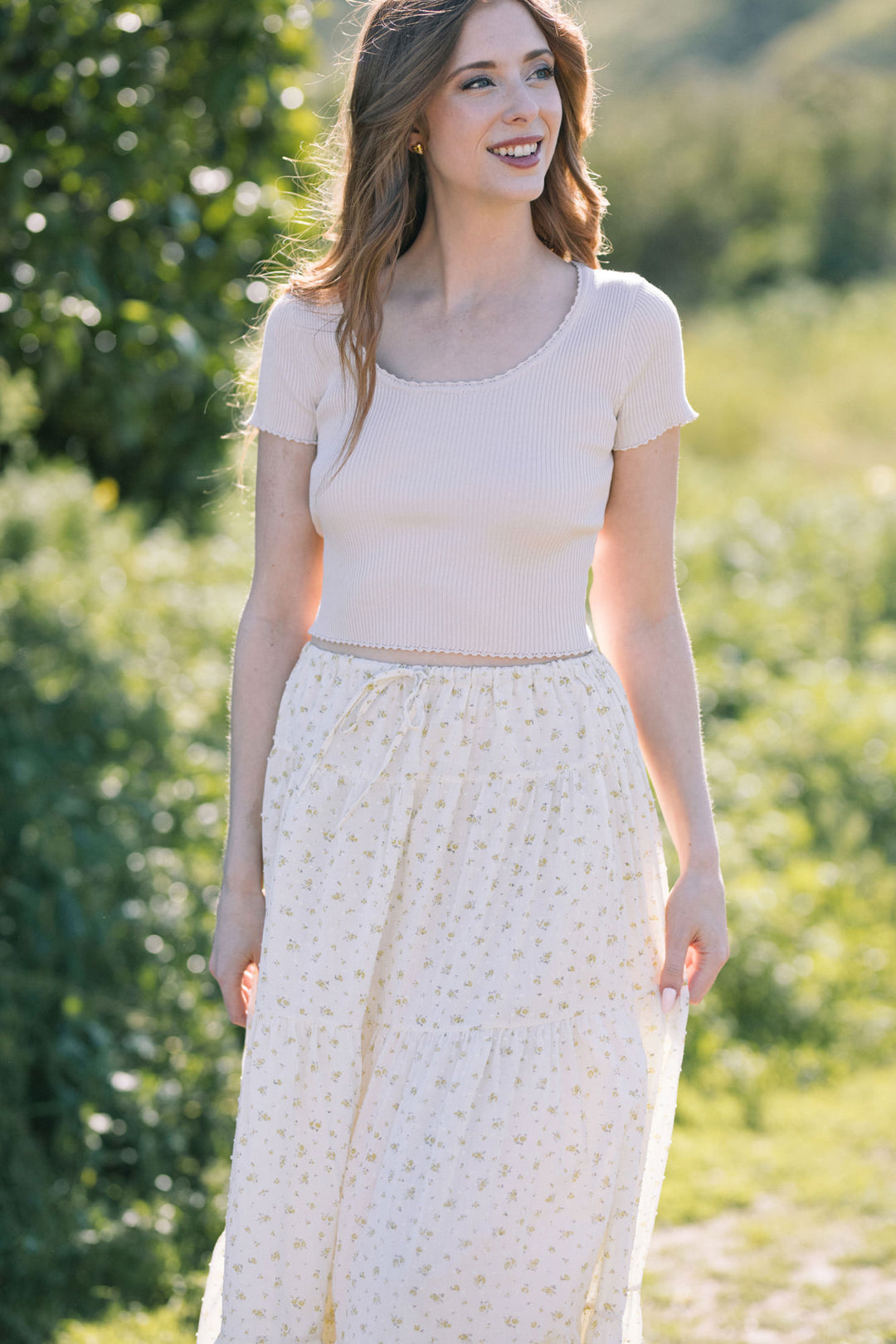 A young woman with long brown hair smiles while wearing a beige ribbed crop top and a white floral-printed midi skirt, standing in a lush, green outdoor setting.