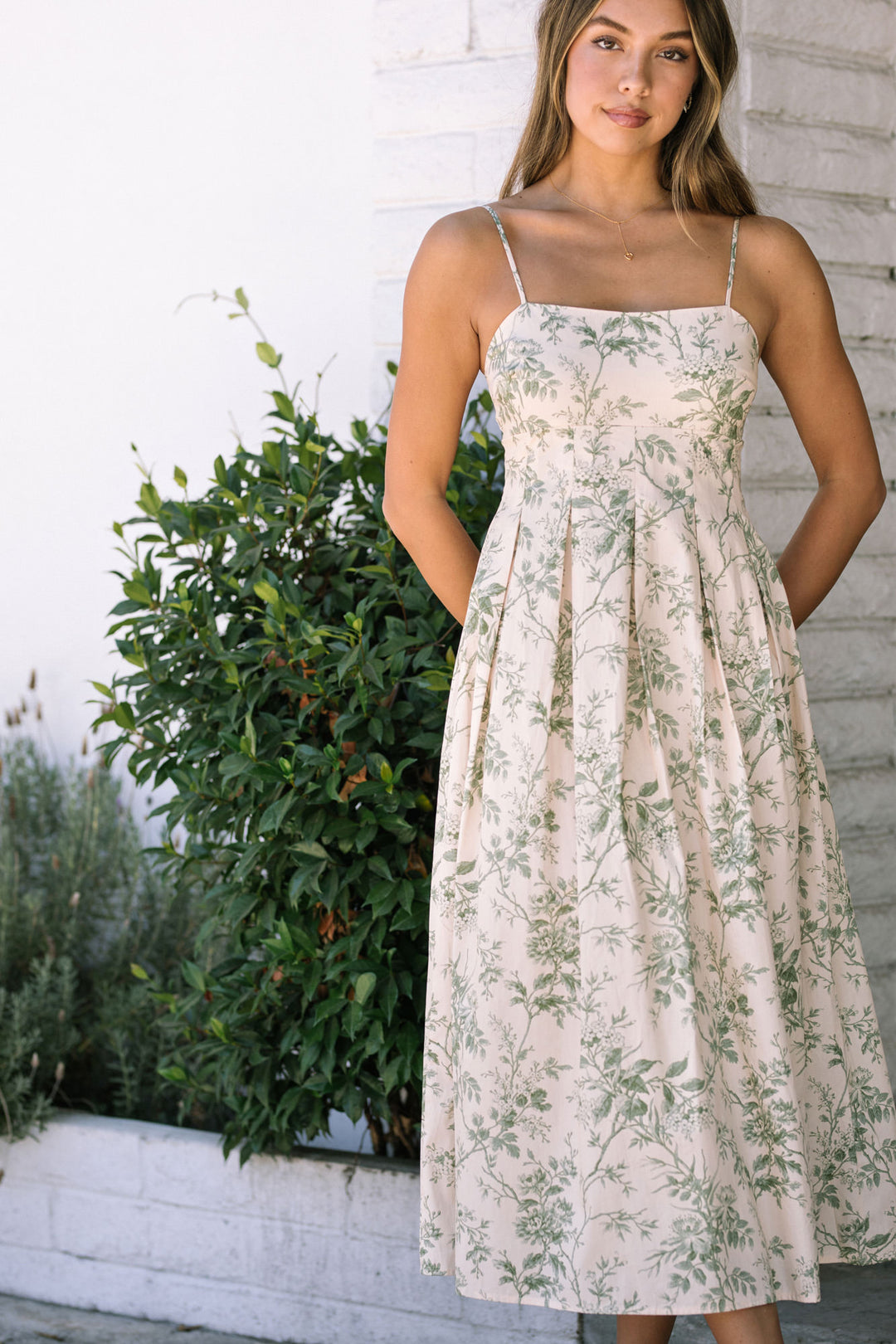 A white midi dress with a v-neck, spaghetti straps, and an allover floral print in shades of green. The dress has a fitted waist and a flowing, ankle-length skirt.