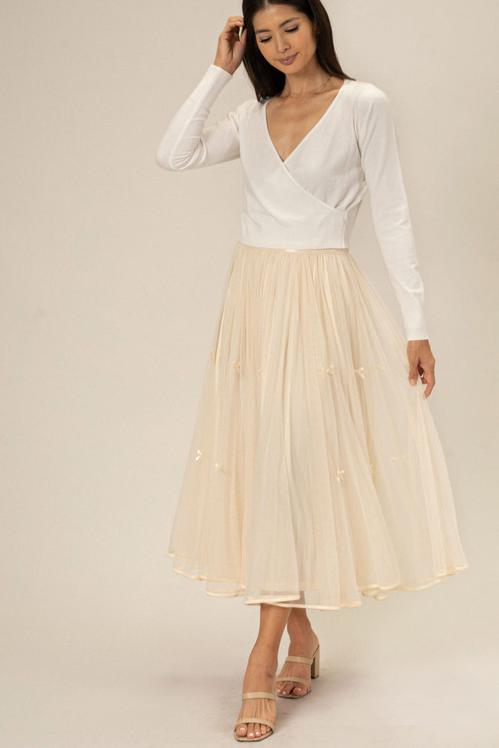 A woman wearing a white wrap top and a long, pleated beige skirt with small bows cascading down the front.