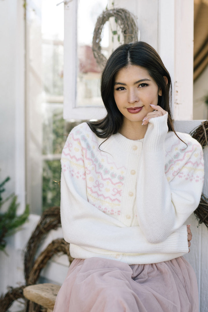 The image shows a young woman wearing a white knit cardigan with a delicate floral pattern. She has long dark hair and is smiling at the camera, standing against a backdrop of a rustic wooden wall.