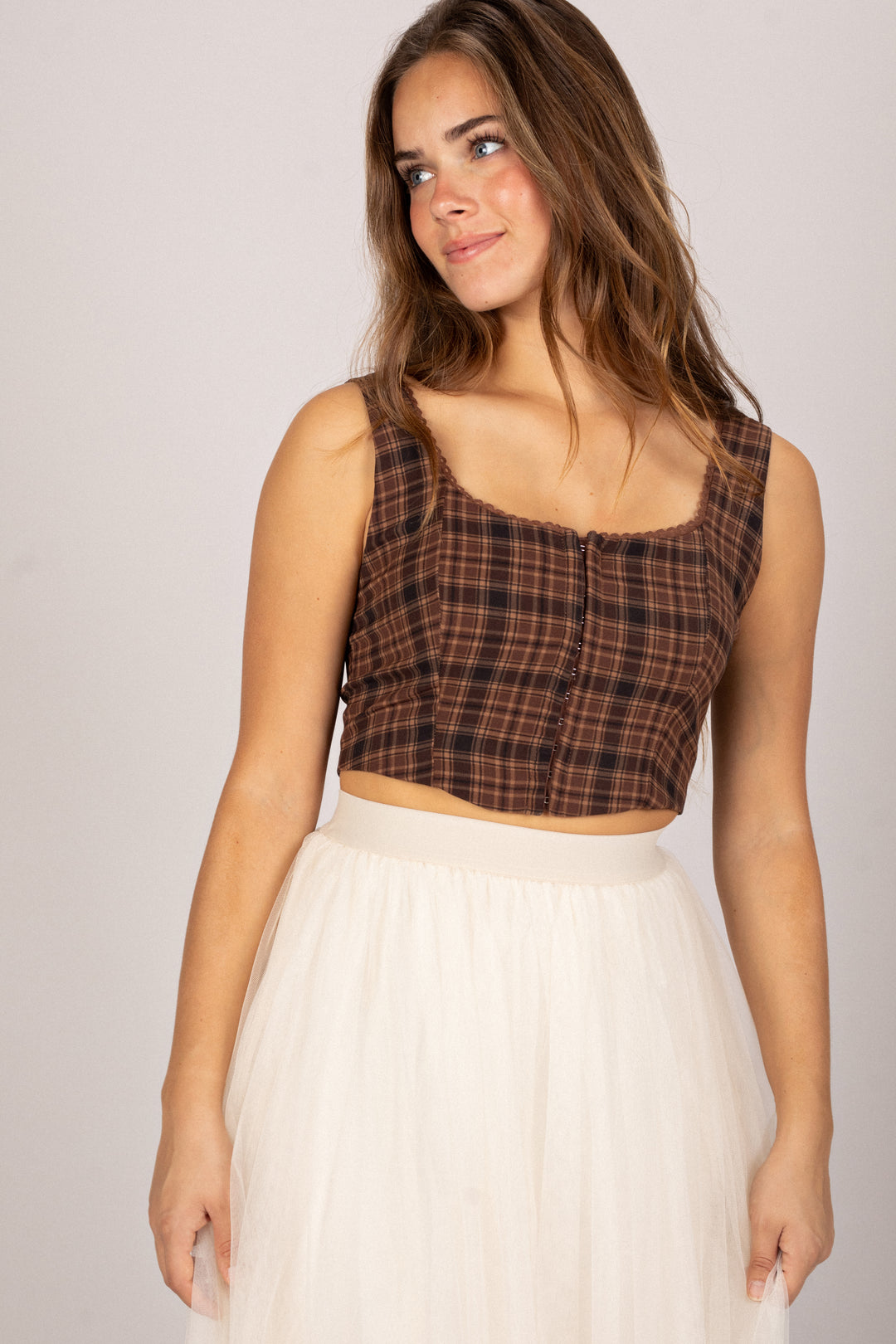 A young woman with wavy brown hair wearing a brown and black plaid crop top and a white skirt, posed against a plain background.