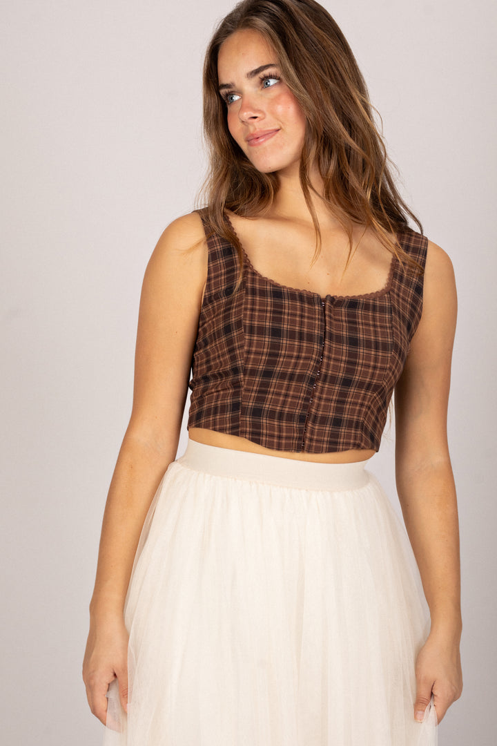 A young woman with wavy brown hair wearing a brown and black plaid crop top and a white skirt, posed against a plain background.