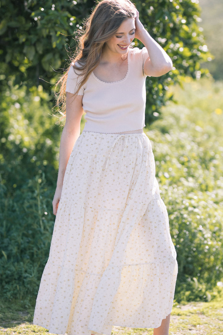 A young woman with long, wavy brown hair is smiling and holding her hair back, wearing a beige knit crop top and a flowing white skirt with yellow floral print.