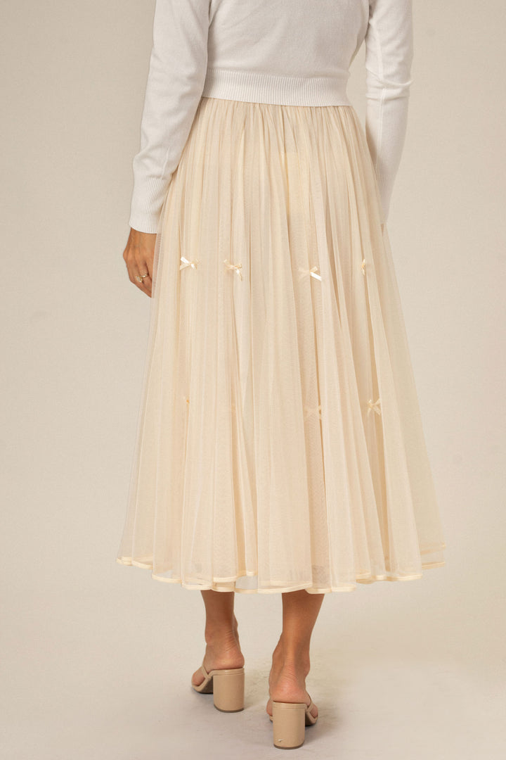 A long, flowy skirt with a tulle overlay, featuring small bows along the hem, in a creamy beige color.