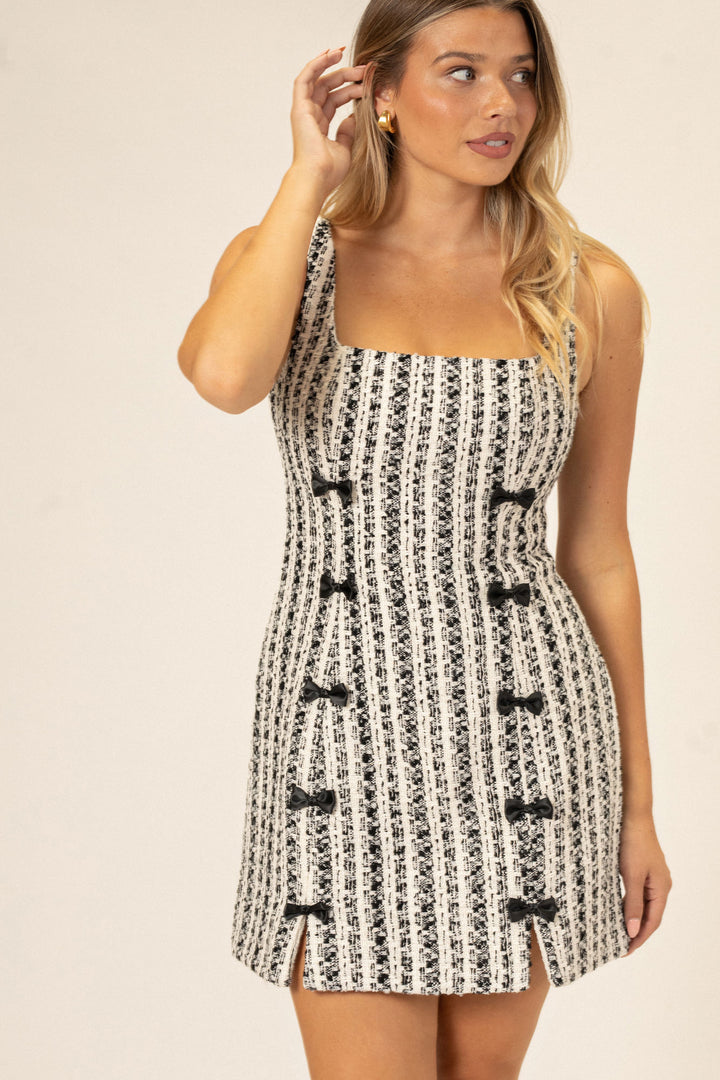 A sleeveless mini dress featuring a fitted, striped tweed pattern in black and white with decorative bow details at the front.