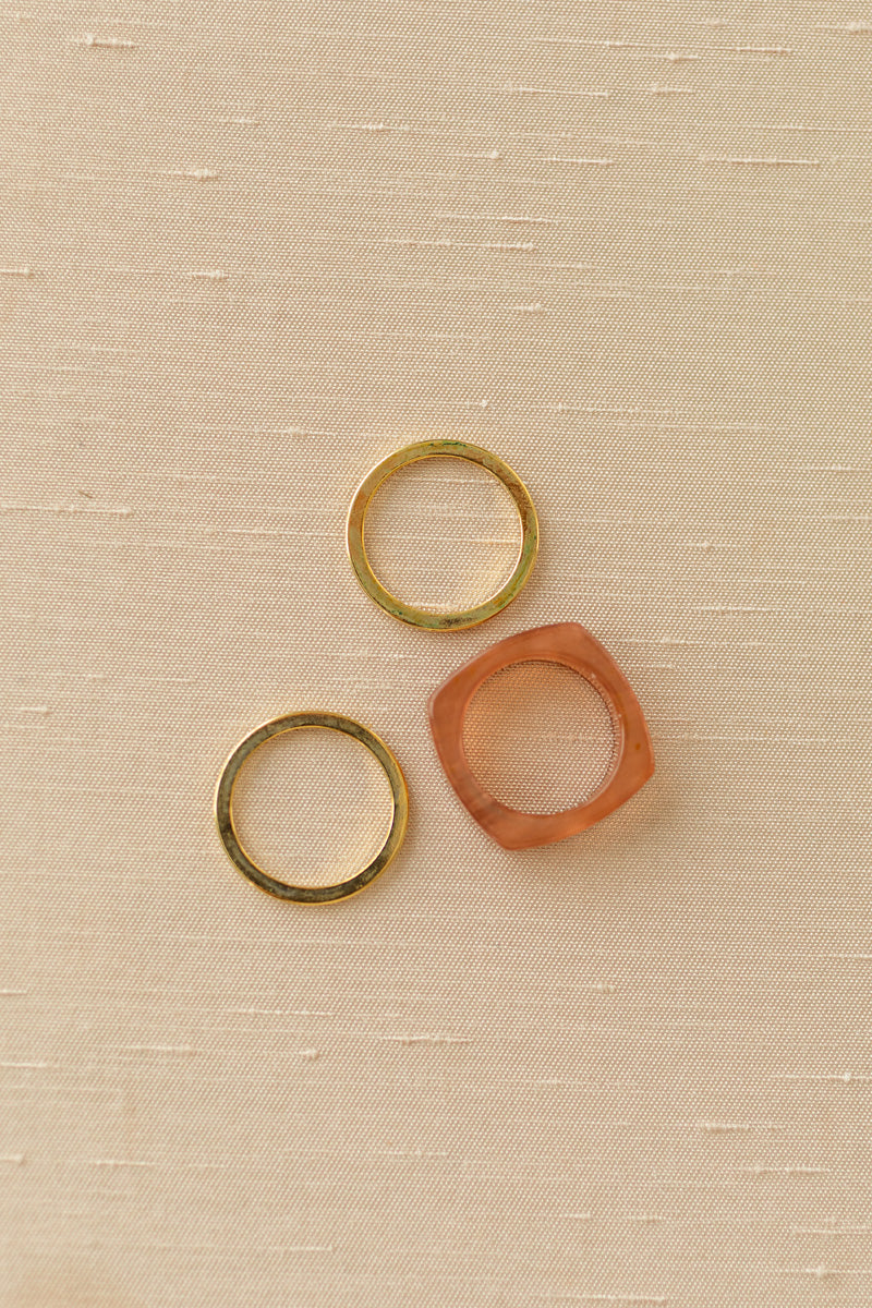 Three stacked rings in shades of gold and coral on a beige fabric background.