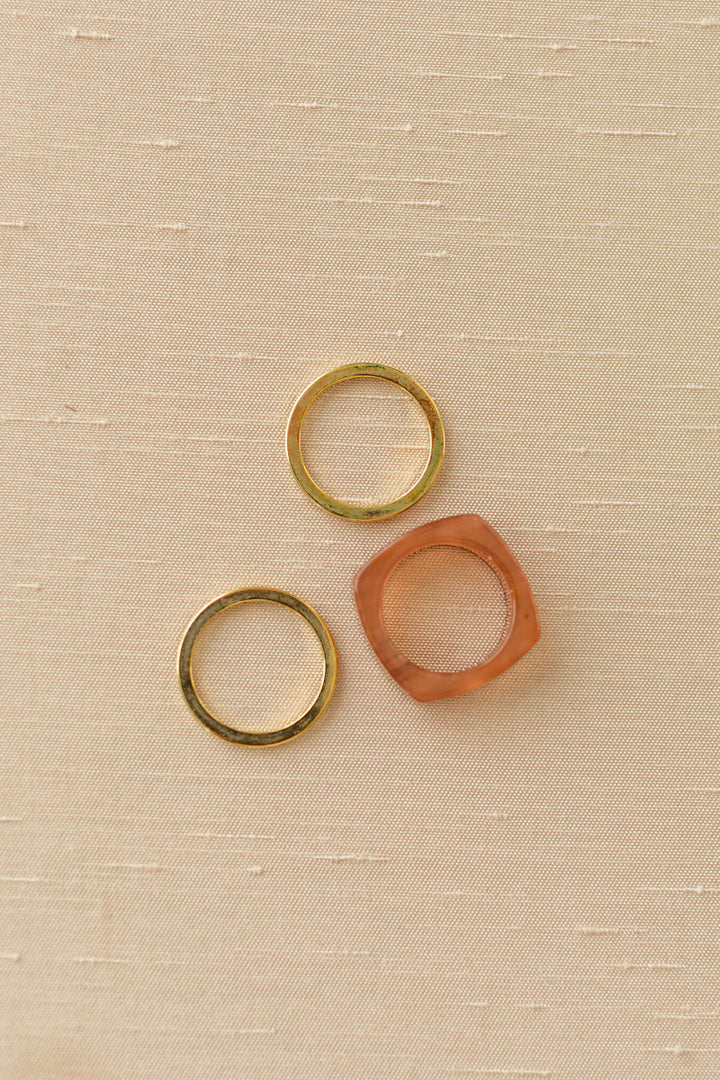 Three stacked rings in shades of gold and coral on a beige fabric background.