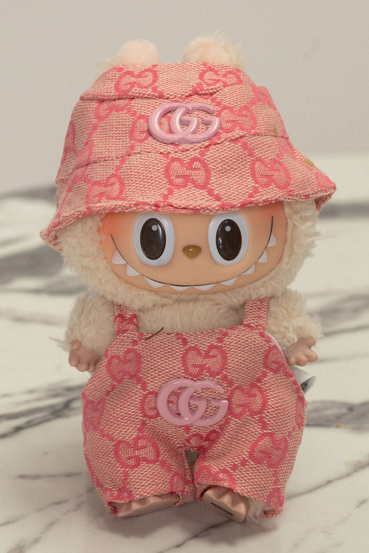 Plush, pink Labubu doll wearing a Gucci-print hat and matching overalls with Gucci logos.