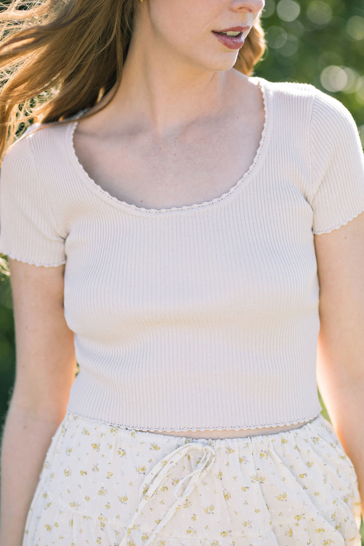 White ribbed crop top with scalloped neckline, worn by a woman with long brown hair standing in a natural setting.