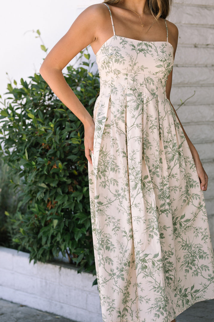 The Alondra Floral Midi Dress features a delicate floral print on a flowing, pleated skirt with spaghetti straps. The dress is styled against a lush, greenery-filled backdrop.