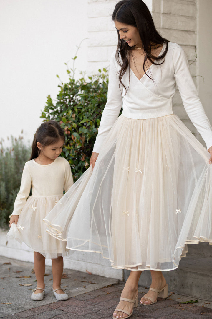 A woman in a white knit top and a long tulle skirt with bows walks beside a young girl wearing a cream-colored dress. The skirt has a flowing, ethereal appearance.