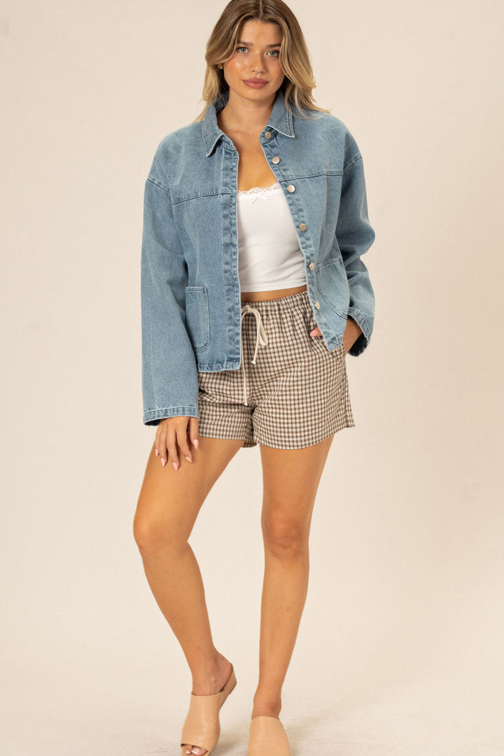 A young woman wearing a denim jacket and plaid drawstring shorts, posing against a beige background.