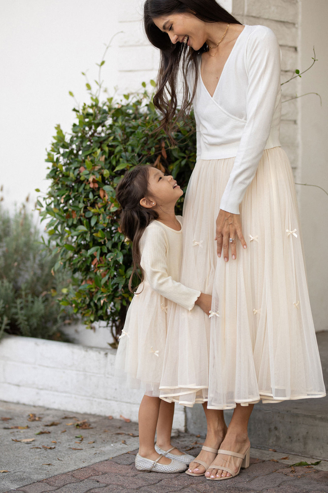 A woman in a white long-sleeved dress and a young girl in a white dress stand together in front of a green shrub, both with long dark hair.