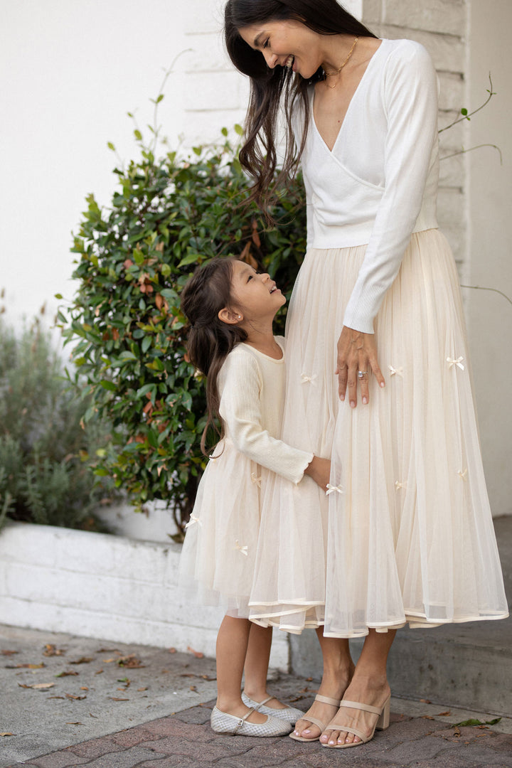A woman in a white long-sleeved dress and a young girl in a white dress stand together in front of a green shrub, both with long dark hair.