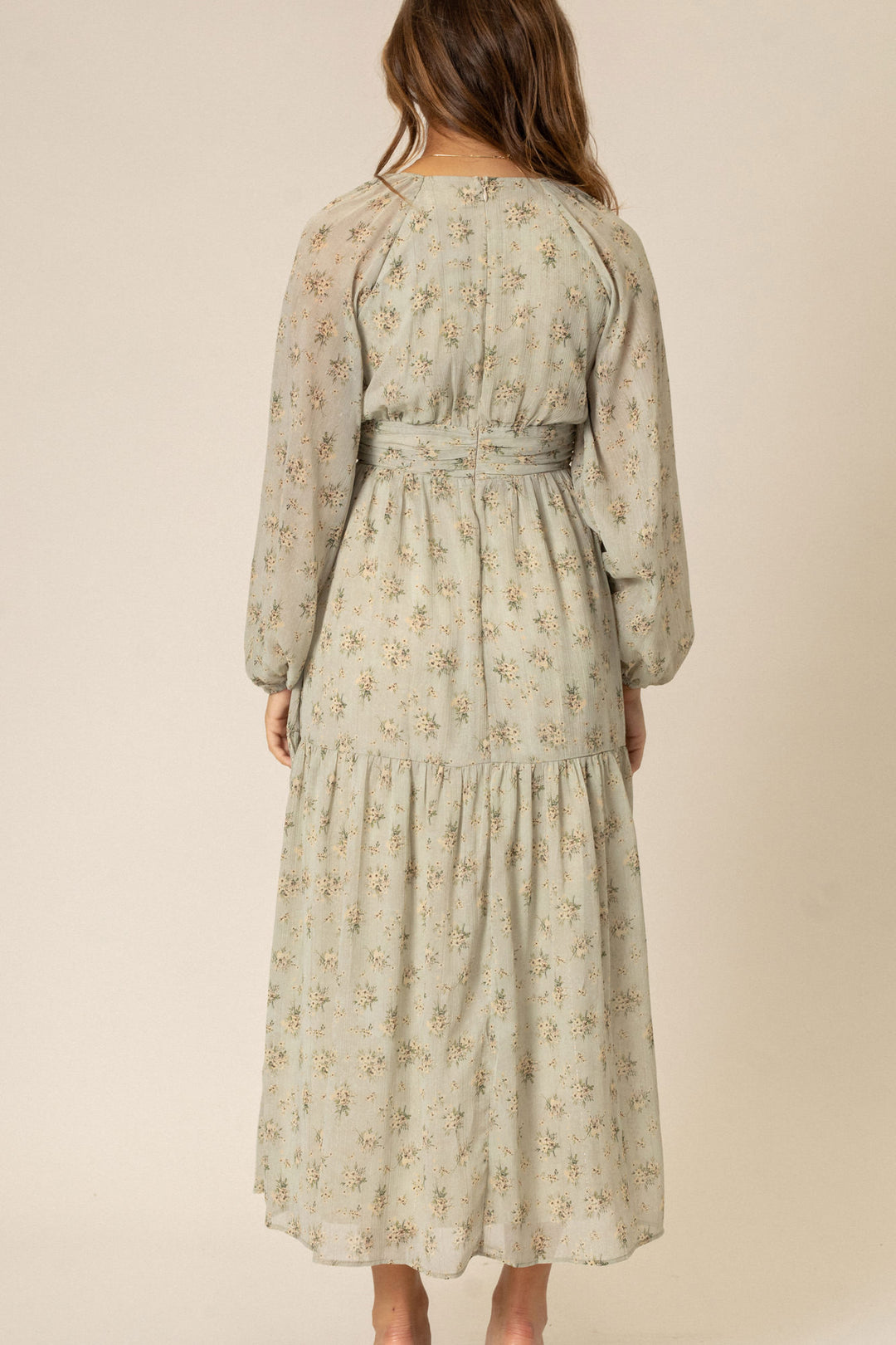 A flowing, long-sleeved floral dress in a soft beige color with delicate floral patterns, featuring a high waist and tiered skirt.