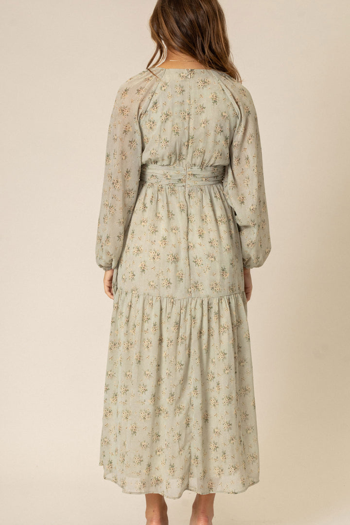 A flowing, long-sleeved floral dress in a soft beige color with delicate floral patterns, featuring a high waist and tiered skirt.