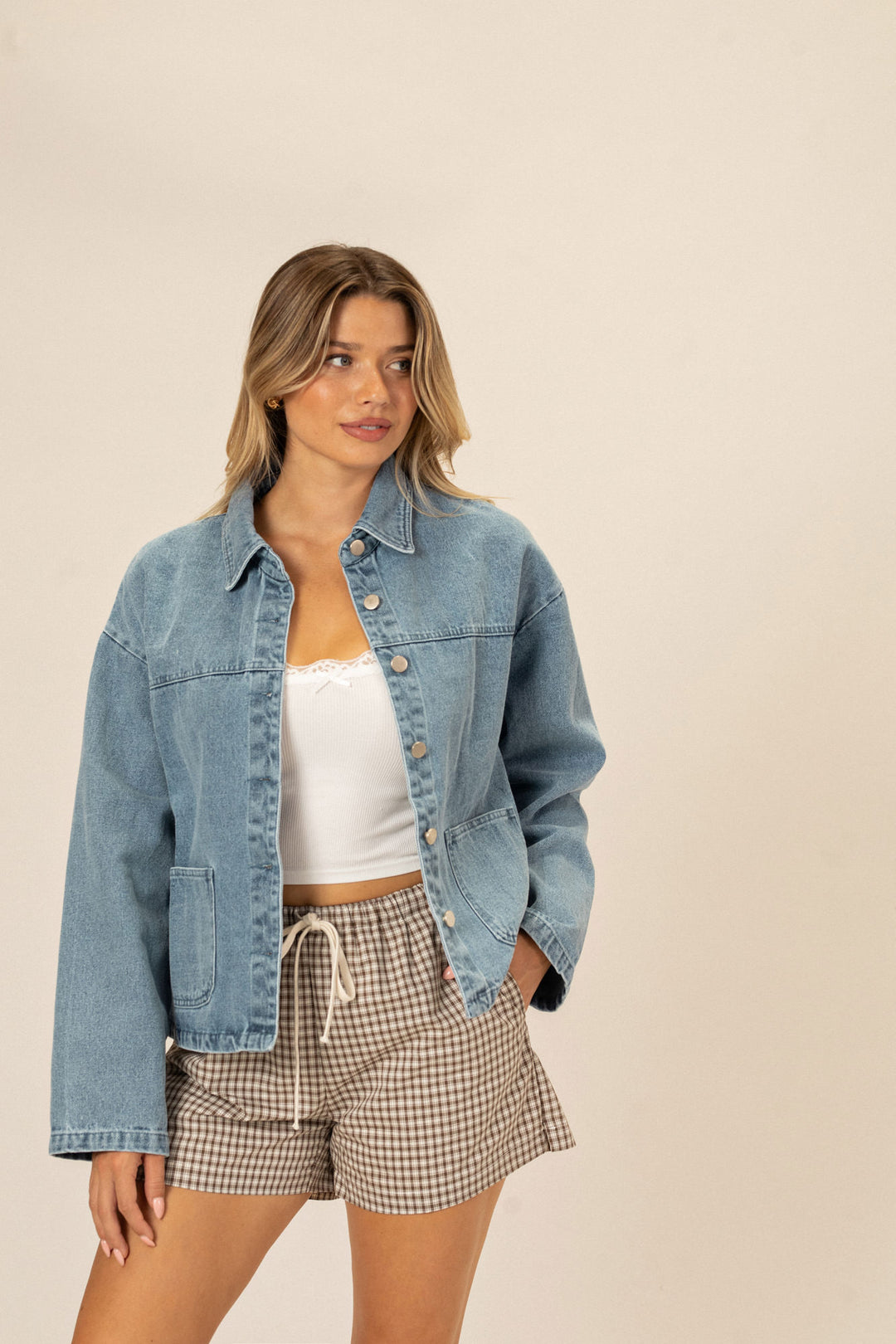 A young woman in a denim jacket and checkered shorts poses against a neutral background. The outfit appears casual and stylish.