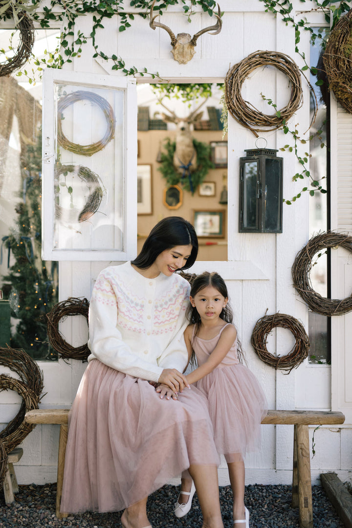 A woman in a white patterned sweater and a young girl in a pink tulle skirt sitting on a wooden bench in front of a rustic white wall decorated with grapevines, woven wreaths, and other natural elements.